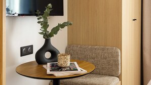 Una habitación moderna con un televisor de pantalla plana montado en la pared, una pequeña mesa redonda con un jarrón y una revista, y un banco acolchado.
