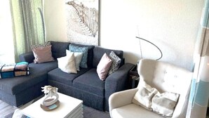 Un salon avec un canapé d’angle gris foncé, un fauteuil blanc et une table basse. Un tableau représentant une clôture et un paysage est accroché au mur.