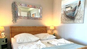 Un lit bien fait, avec du linge de lit blanc et une couverture bleue à motifs, une tête de lit en osier et deux lampes de chevet.