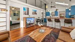A spacious living area with a wooden coffee table, a TV, and a sofa. A kitchen with blue cabinets and a bar area is visible in the background. - Hot Tub Hideout | Old Town’s Best Group Stay (Fort Collins)