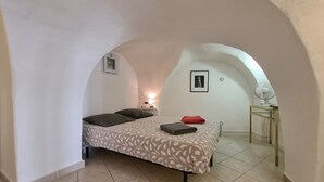 A bedroom with a bed, pillows, a red towel, a fan, and a small table with a lamp. - Gîte Lucas 4 pers. piscine et clim à Sommières - Calm, nature & traditions ! (Sommières)