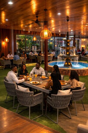A restaurant with a dining area featuring round tables, wicker chairs, and a central water fountain. - Golden Crystal Hotel (Accra)