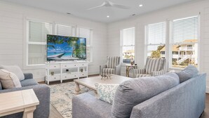 A spacious living room with a large flat-screen TV, comfortable seating, and large windows with blinds. - Dancing Pelican-New 5-bedroom 3.5 baths, canal house w/elevator, 3min to beach (Holden Beach)