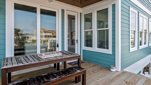 A covered porch with a wooden table and bench, white-framed glass doors, and teal siding. - Dancing Pelican-New 5-bedroom 3.5 baths, canal house w/elevator, 3min to beach (Holden Beach)