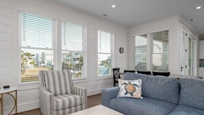 A living room with a sofa, armchair, and coffee table. There are large windows with blinds, a clock on the wall, and a view of buildings outside. - Dancing Pelican-New 5-bedroom 3.5 baths, canal house w/elevator, 3min to beach (Holden Beach)