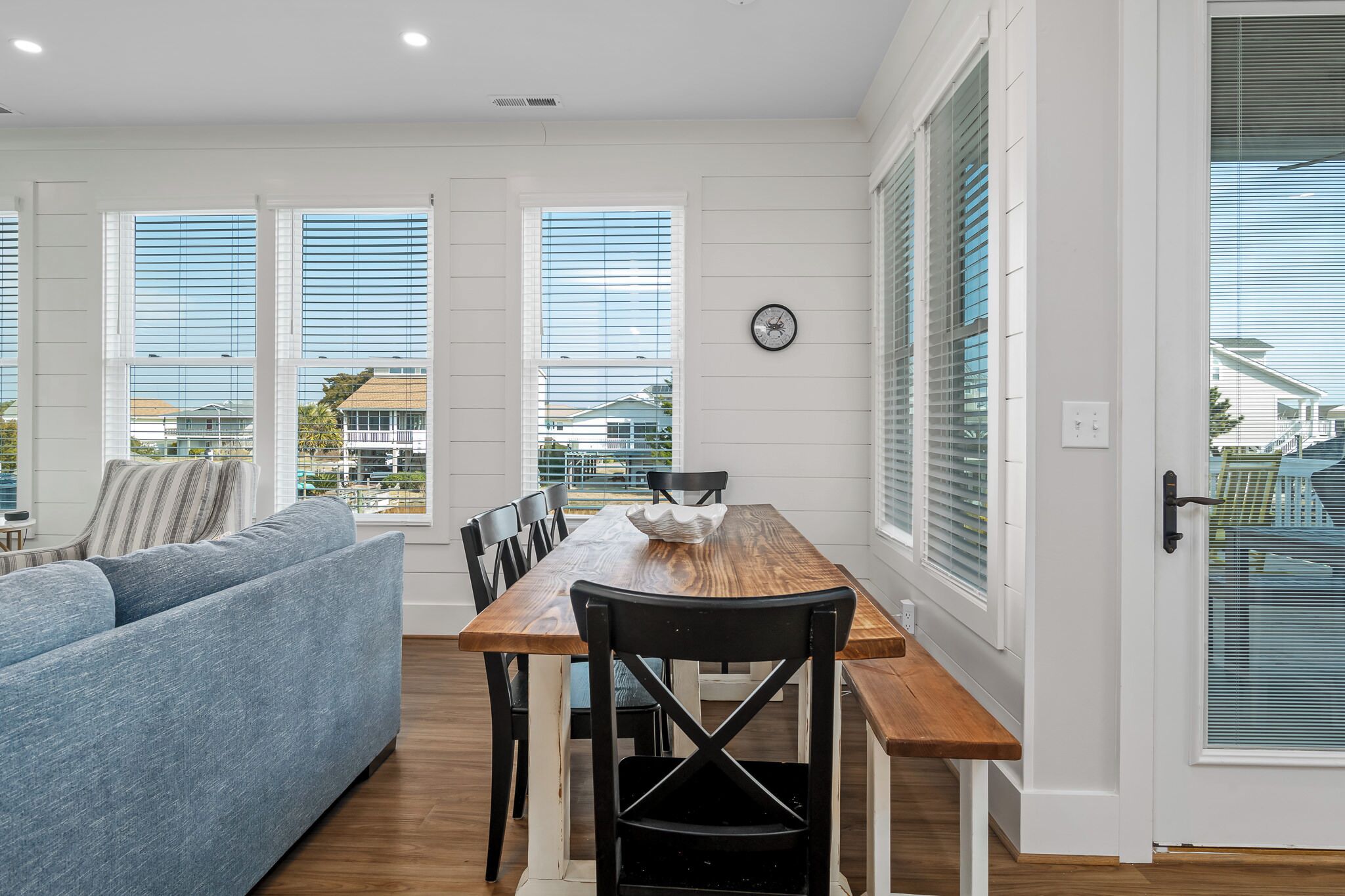 A modern living room with a wooden dining table, blue sofa, and large windows with blinds.