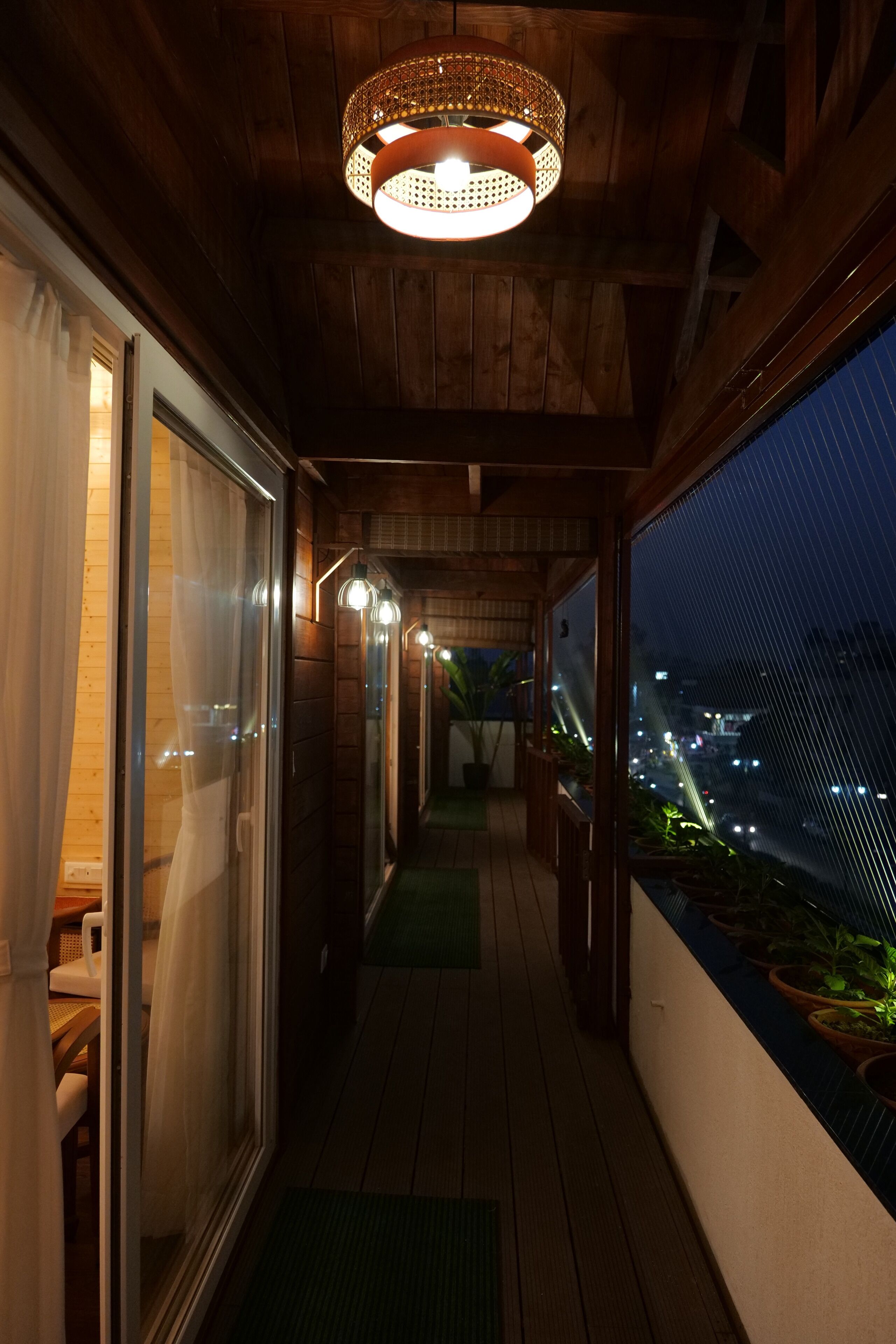 Un pasillo de madera con una lámpara en el techo, vistas de la ciudad por la noche y plantas en macetas a lo largo de la pared.