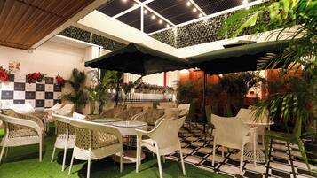 An outdoor seating area with wicker chairs and tables, surrounded by potted plants and a checkered floor pattern.
