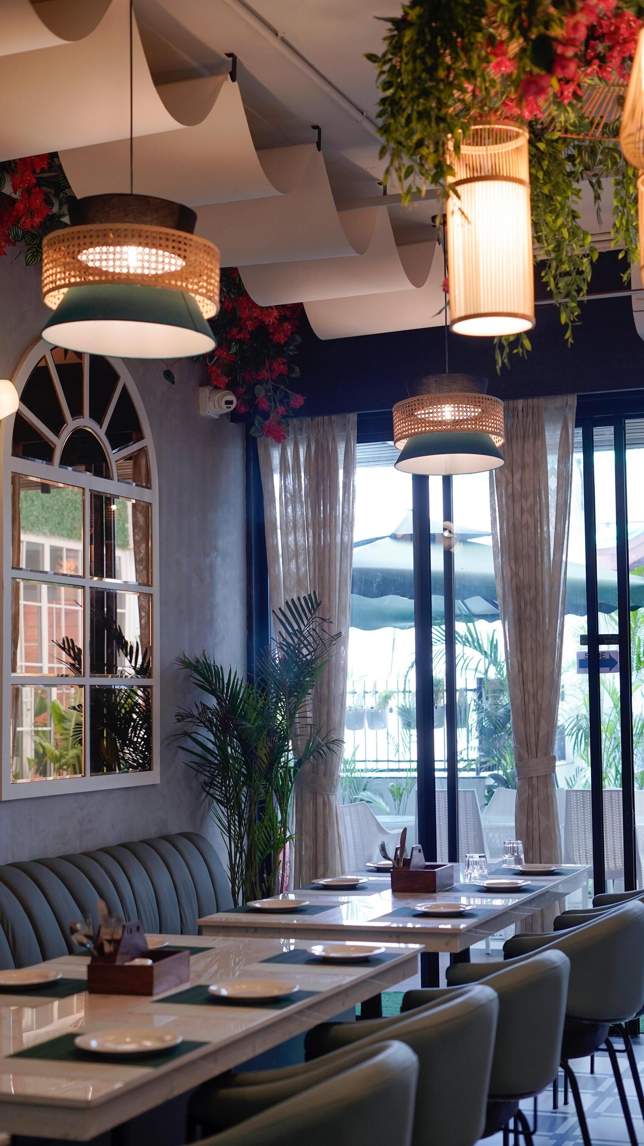 A restaurant with a long counter, cushioned seating, hanging lamps, and large windows with curtains.