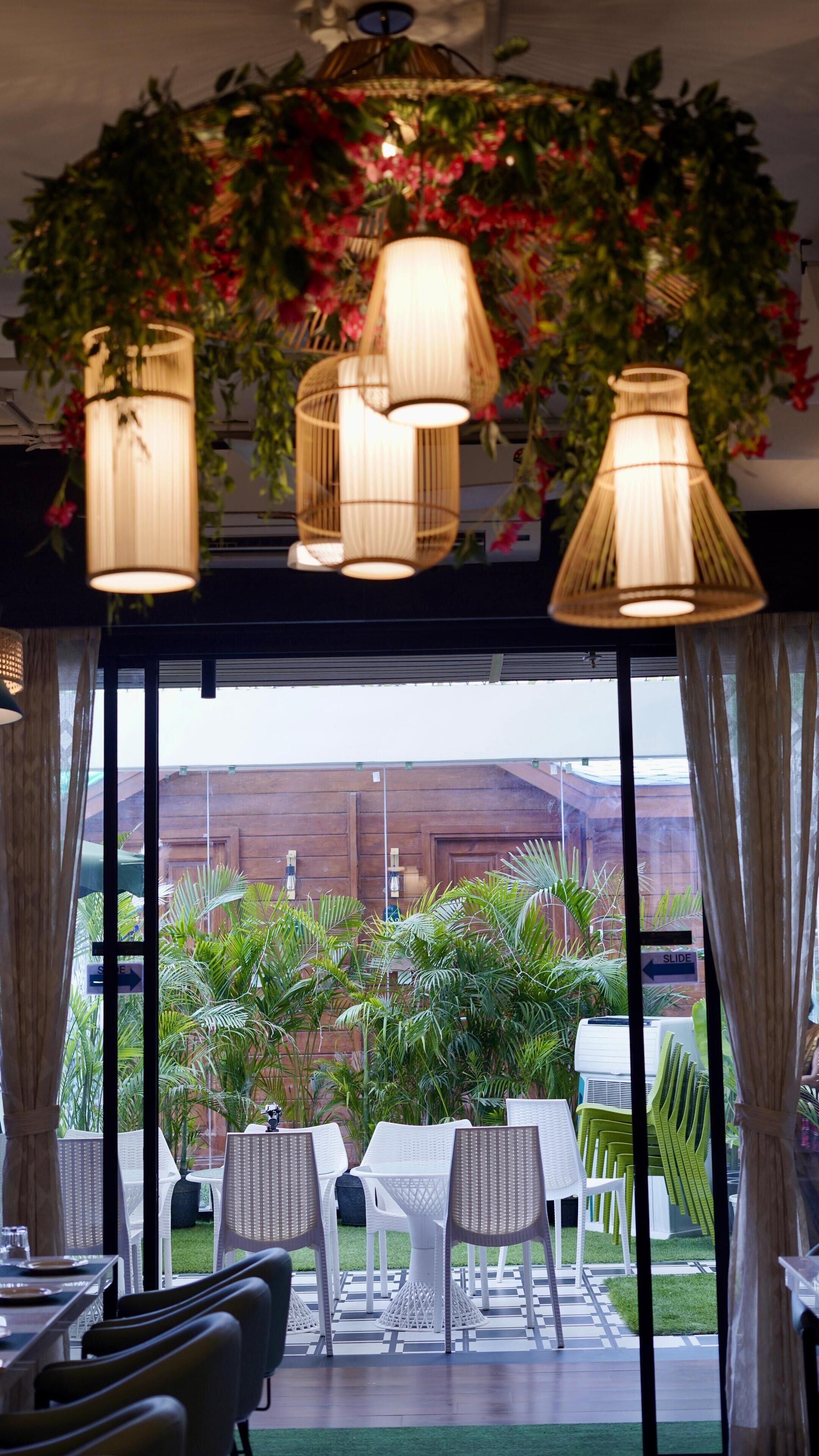 An indoor dining area with wicker chairs and tables, a view of the outdoors through large windows, and hanging lamps with a floral arrangement above.