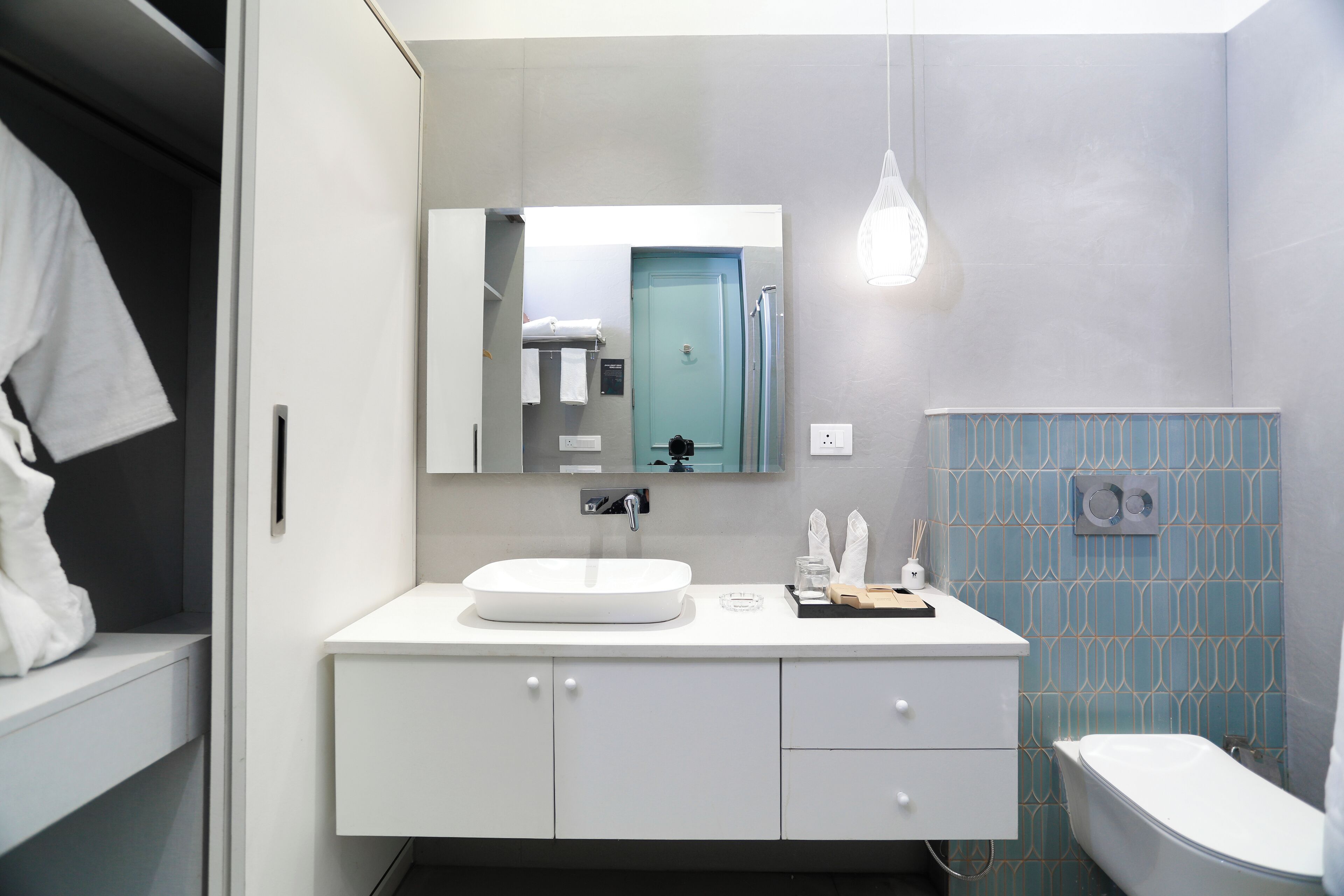 A modern bathroom with a white sink, a mirror above it, and a toilet. There is a towel rack with towels, a small shelf with toiletries, and a light fixture above the mirror.