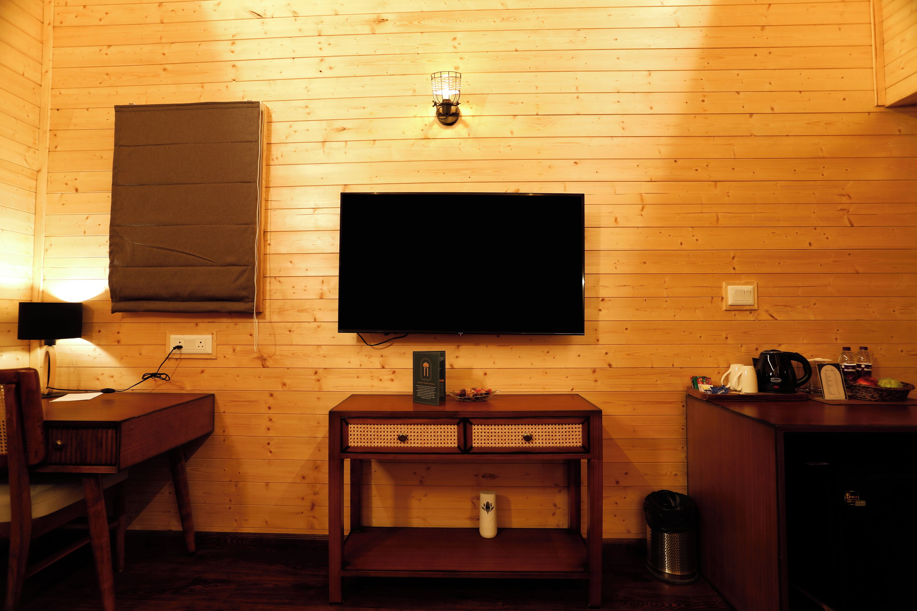 A wooden-paneled room with a television mounted on the wall, a desk with a lamp, and a small cabinet with various items on top.