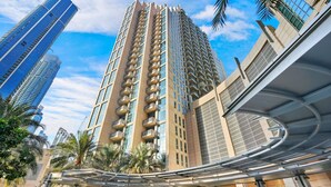 A high-rise building with a modern design, featuring a curved pedestrian bridge and surrounding palm trees.