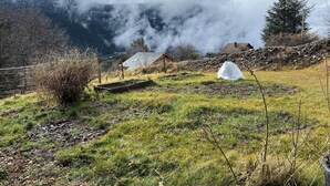 Paisagem rural com muro de pedra, construção de madeira e uma casa ao fundo.