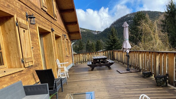 Um deck de madeira com móveis de exterior, incluindo mesa e cadeiras, com vista para uma paisagem montanhosa.