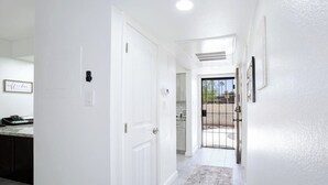 Un pasillo con una puerta blanca, una cocina visible a través de una puerta abierta y una pequeña alfombra en el suelo.