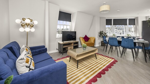 A modern living room with a blue sofa, a coffee table, and a dining area with blue chairs. - Buzzing Brighton Apartments | By My Getaways (Brighton)