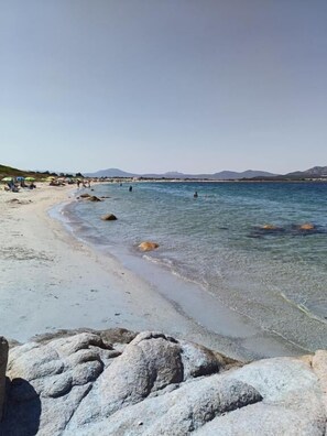 A beach with clear water, rocks, and distant mountains. - Studio's Pavese (Olbia)