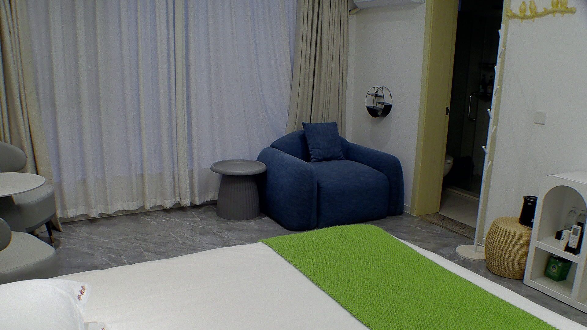 A hotel room with a bed, a blue armchair, a small round table, a green rug, and a white shelf with items.