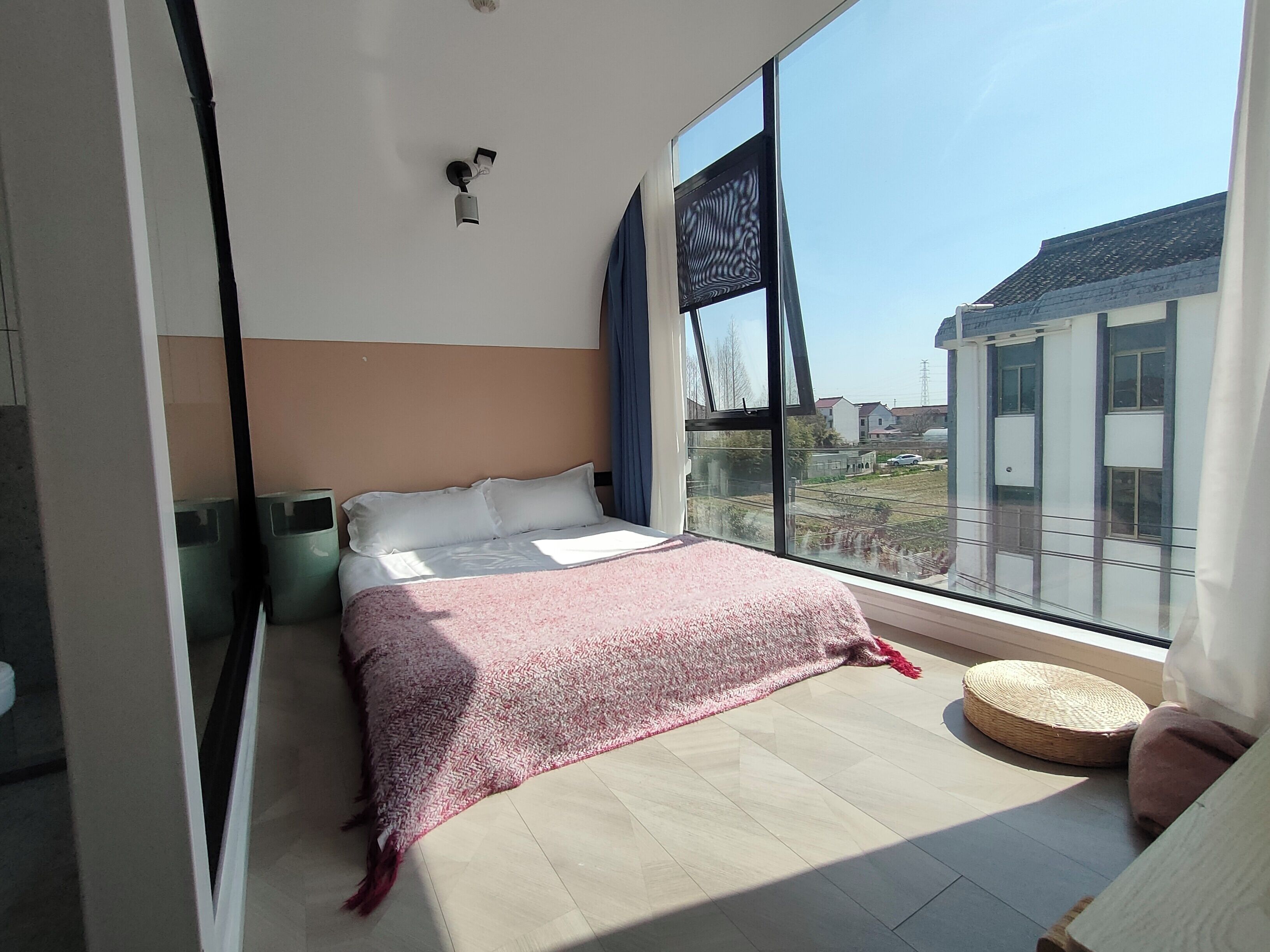 A modern bedroom with a large window, a bed with a pink blanket, a small round table, and a wall-mounted light fixture.