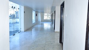 A long, narrow hallway with white walls and a marble floor. There are large windows on one side, allowing natural light to enter. - HOTEL TRIDEV  (Reasi)