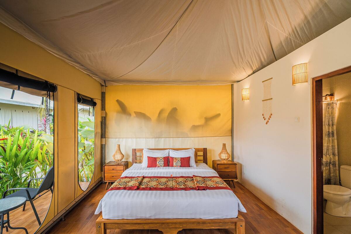 Un dormitorio con cama, mesitas de noche, una ventana con vista a follaje y una puerta que da al baño.