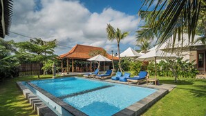 A swimming pool with lounge chairs and umbrellas, surrounded by greenery and a building with a tiled roof. - Paddy's Glamping (Munggu)