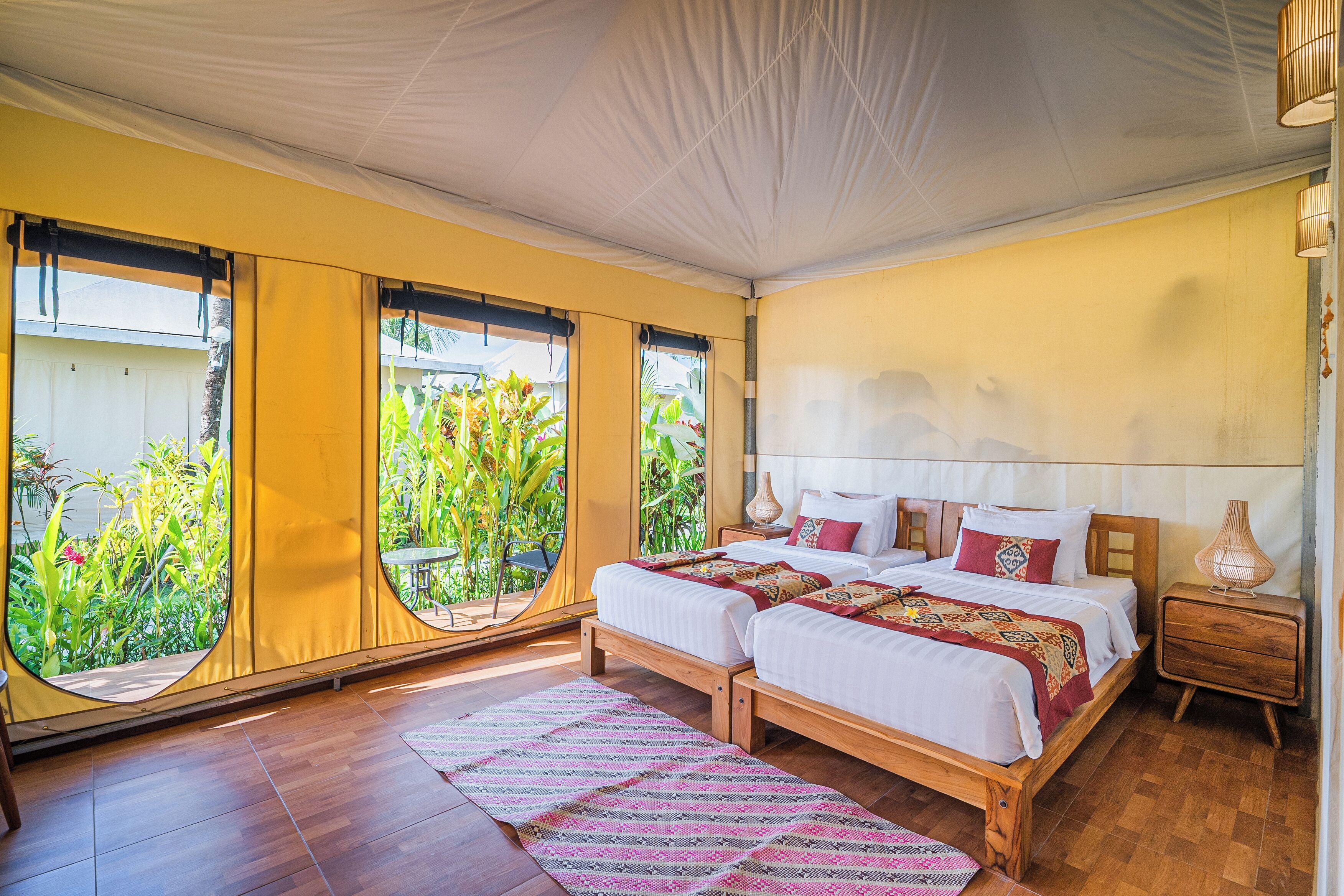 A room with two beds, a wooden floor, a large window with a view of greenery, and a bedside table with a lamp.