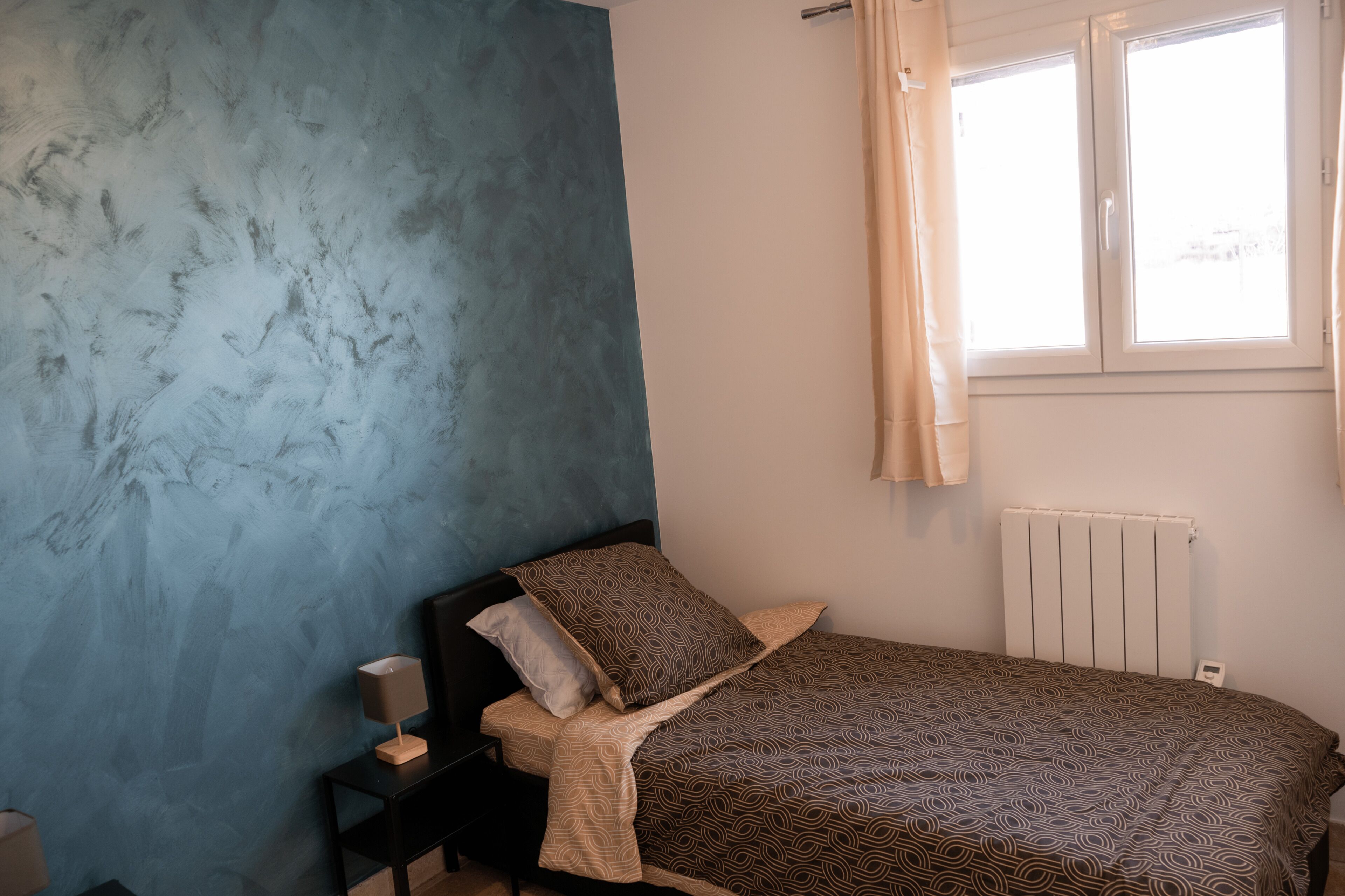 Ein Schlafzimmer mit einem Bett, einem Nachttisch, einer Lampe, einem Fenster mit Vorhängen und einer Heizung.