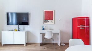 Una habitación moderna con un escritorio blanco, un armario rojo, una silla blanca, un televisor y un cartel enmarcado en la pared.