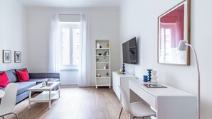 Una sala de estar moderna con un sofá gris, una mesa de centro blanca y un escritorio blanco con una computadora. Hay una estantería con objetos decorativos y un cuadro grande enmarcado en la pared.