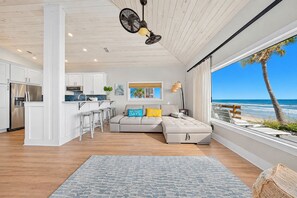 A modern living space with a kitchen, a sofa with cushions, a dining area, and a large window with a view of the ocean.