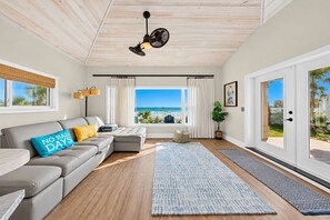 A modern living room with a view of the beach, a grey sofa, a blue rug, and a ceiling fan.