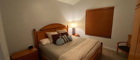 Ein Schlafzimmer mit einem hölzernen Bett, einem Nachttisch, einer Lampe, einem Fenster mit Jalousien und einem Deckenventilator.