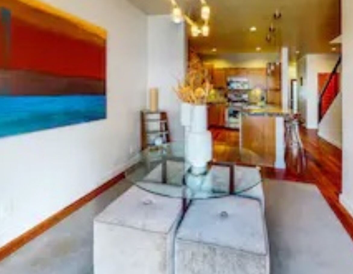 Un interior moderno con una mesa de centro de vidrio, un cuadro abstracto rojo y una zona de cocina al fondo.