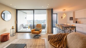 A modern living room with a sofa, a coffee table, and a dining area with a table and chairs. There is a large window providing a view of the outdoors. - Apartment in Nieuwpoort With Balcony (Nieuwpoort)