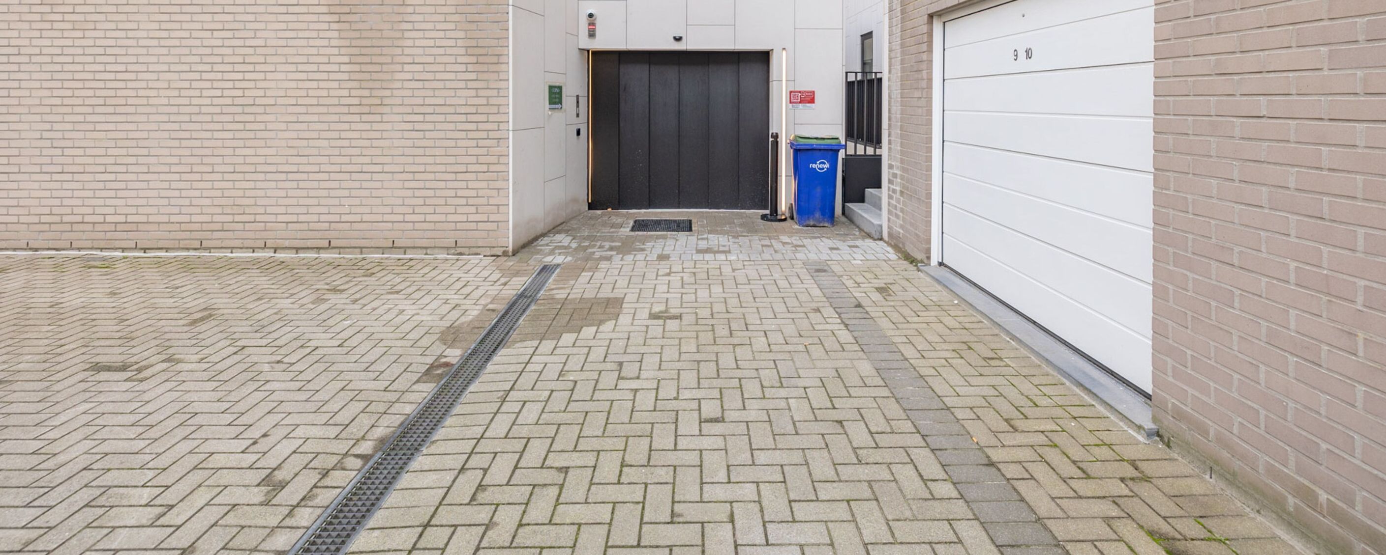 A narrow brick corridor with a metal grate on the floor, a closed door, and a blue trash bin.