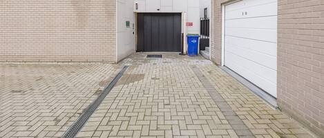 A narrow brick corridor with a metal grate on the floor, a closed door, and a blue trash bin.