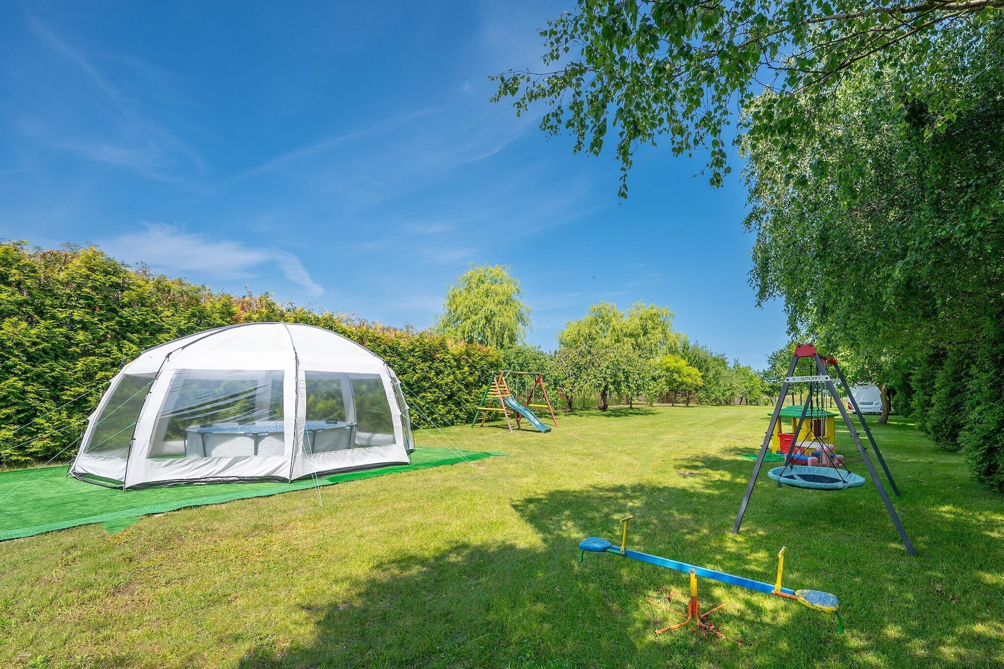 Ein Zelt, eine Schaukel und ein Trampolin auf einer grünen Wiese mit Bäumen.