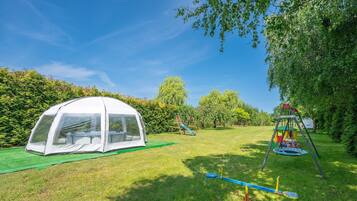 Ein Zelt, eine Schaukel und ein Trampolin auf einer grünen Wiese mit Bäumen.