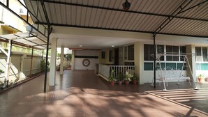 An indoor covered area with a tiled floor, potted plants, a metal swing, and a view of a building with a garage door in the background. - Heritage Garden Villa (Mangaluru)