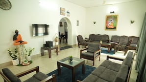 A living room with a Buddha statue, grey sofas, a TV, and a dining area in the background. - Heritage Garden Villa (Mangaluru)