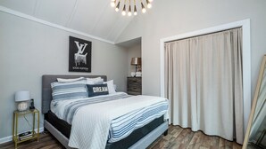 A bedroom with a bed, a bedside table with a lamp, a framed picture on the wall, and a window with curtains. - Three Oaks Unit Steps To Downtown! 2 Night Summer Min! (Three Oaks)