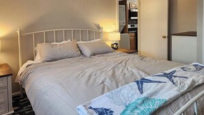 Un lit avec une couette et des oreillers de couleur beige, une table de chevet avec une lampe, et une vue sur un coin cuisine et une salle de bain.