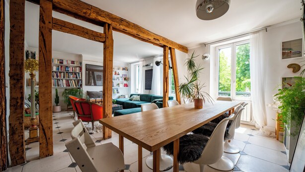 Un coin repas avec une table et des chaises en bois, une bibliothèque et une vue sur un salon comprenant un canapé et une plante.