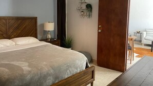 Un dormitorio con una cama de madera, una lámpara de noche, una mesita con lámpara, una planta en maceta y una puerta de madera.