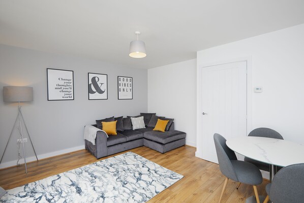 A modern living room with a grey sectional sofa, a marble-patterned rug, a white dining table with grey chairs, and a floor lamp. - 3 Bedroom Large House - Train Station (Derby)