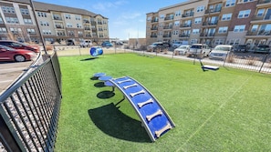 A playground with a slide and swings, surrounded by a fence, with apartment buildings in the background.