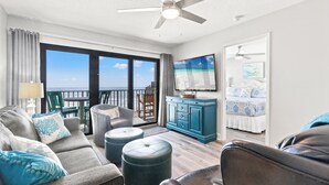 Un salon moderne avec un canapé, des poufs, un téléviseur et un balcon offrant une vue sur l’océan.
