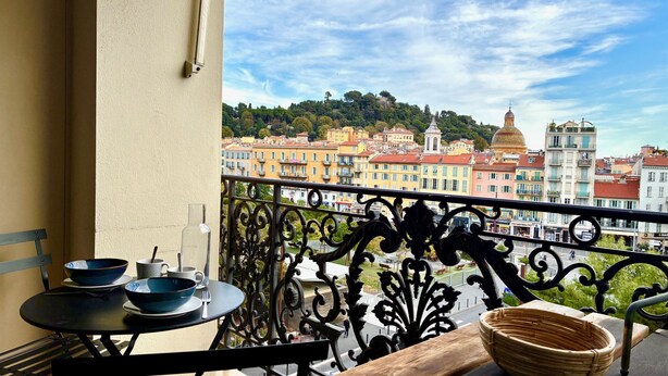 A balcony with a view of colorful buildings, a table with bowls and a bottle, and a decorative metal railing. - Terrasse Felix Faure (Niza)